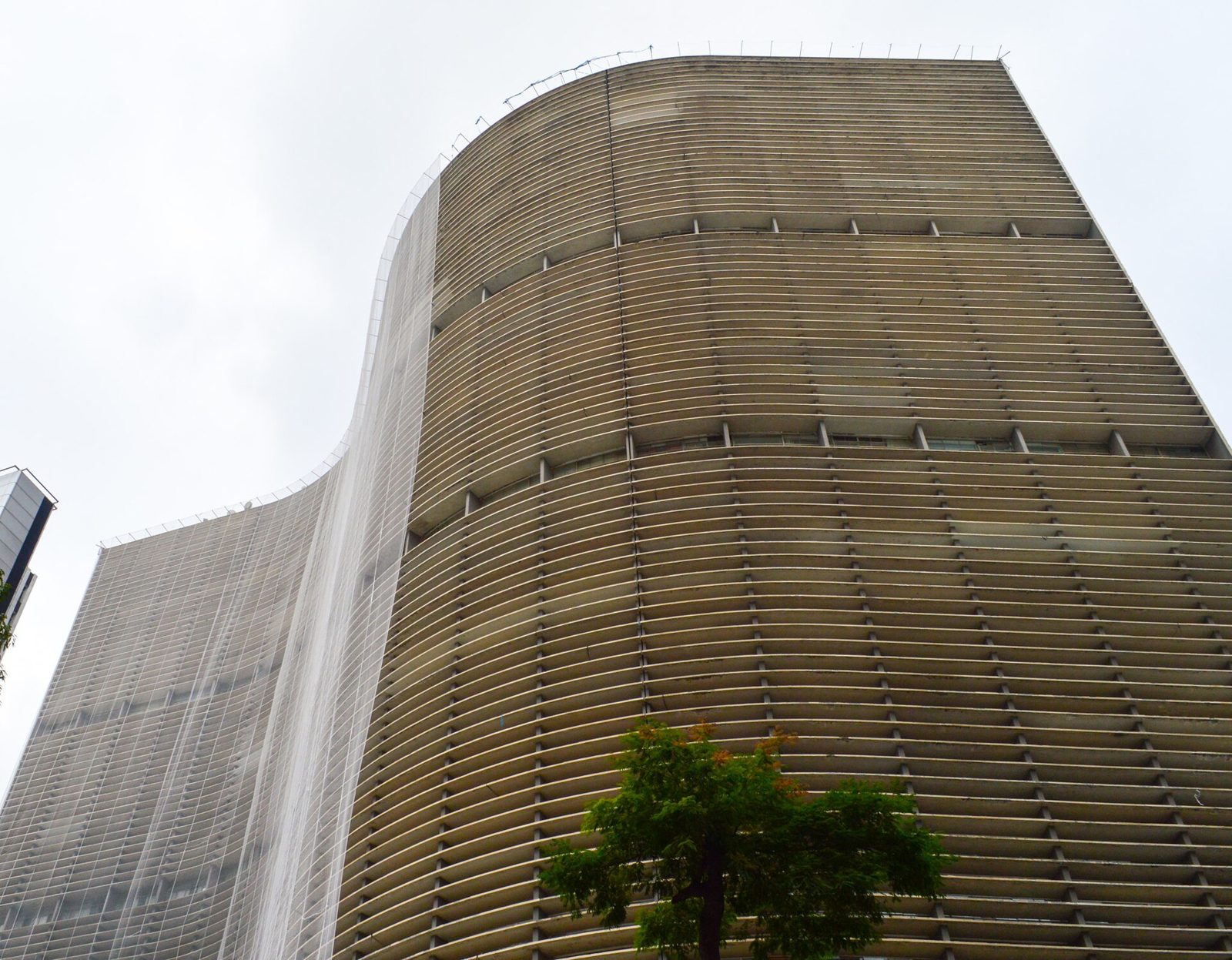 A lâmina sinuosa vista de baixo, ainda coberta pela tela: os brises horizontais de concreto desenham a fachada norte